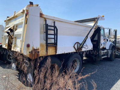 Dump truck bed showing hydraulic system and rear dual tires with rusted edge and ladder on side.
