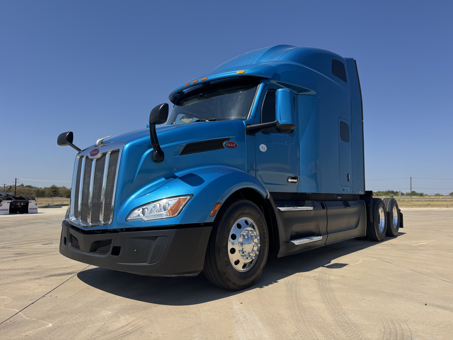 Peterbilt 579 conventional sleeper truck in blue, front-left view, showing cab, grille, large chrome wheels, side mirrors, and aerodynamic design.