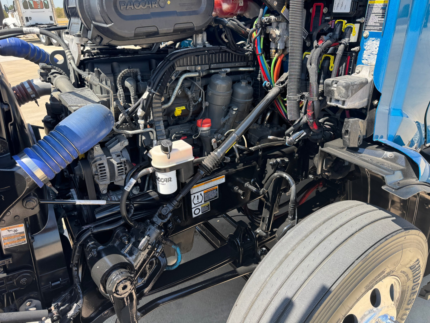 Peterbilt 579 engine bay showing diesel engine, various hoses, wiring harnesses, coolant reservoir, and front axle components.