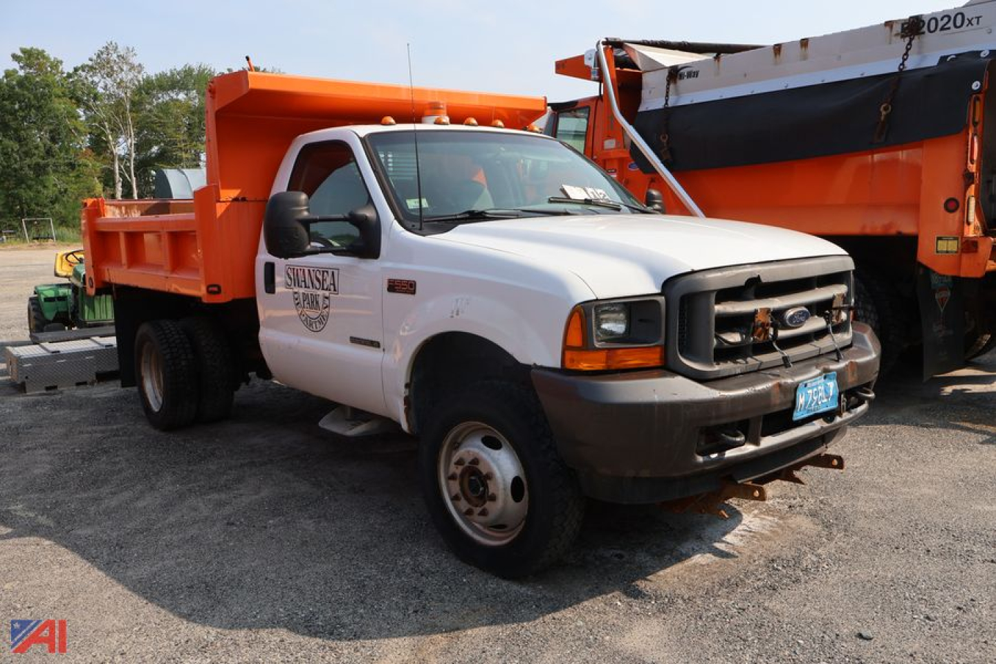 Dump truck front-left view showing white cab, orange dump bed, black grille, and front bumper with blue license plate; Ford emblem on grille, company logo on door.