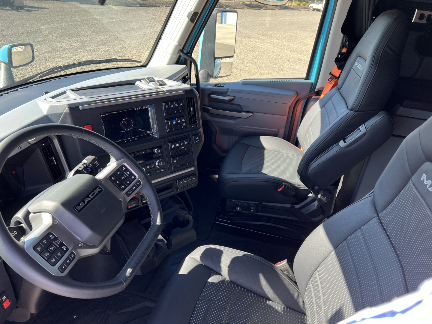 Cargo truck cab interior showing steering wheel with Mack logo, dash with touchscreen, and two grey fabric seats in a Mack PR64T76H truck.