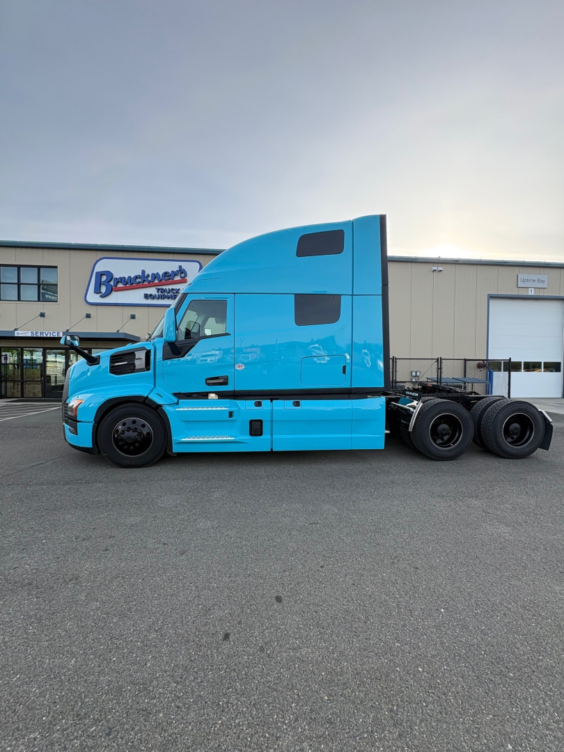 New blue Mack PR64T76H conventional sleeper truck side view, with two rear axles, parked in lot in front of dealership building.
