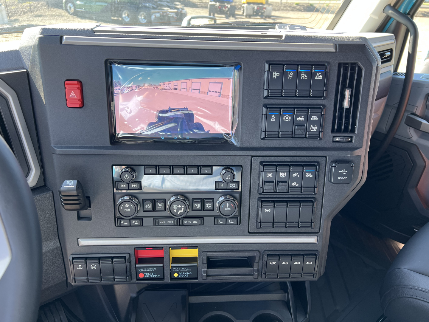 Dashboard of a Mack truck cab showing multiple controls, a screen, and various switches. Visible text not legible, but layout indicates media and climate controls with auxiliary panels.