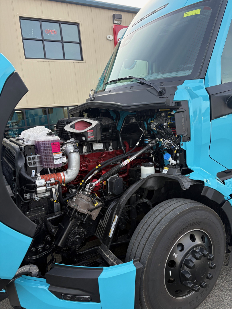 Tractor truck engine bay with open hood showing red engine block and various hoses, wiring, and components in a blue Mack PR64T76H cab.