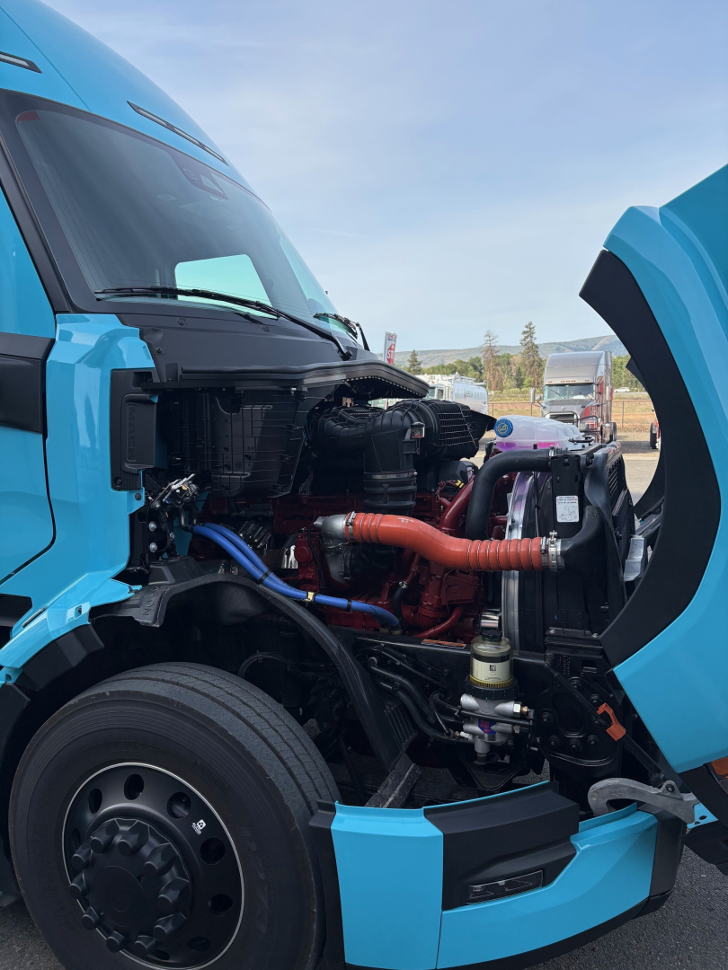 Truck engine bay of a blue Mack PR64T76H with open hood, showing engine components, hoses, and radiator.