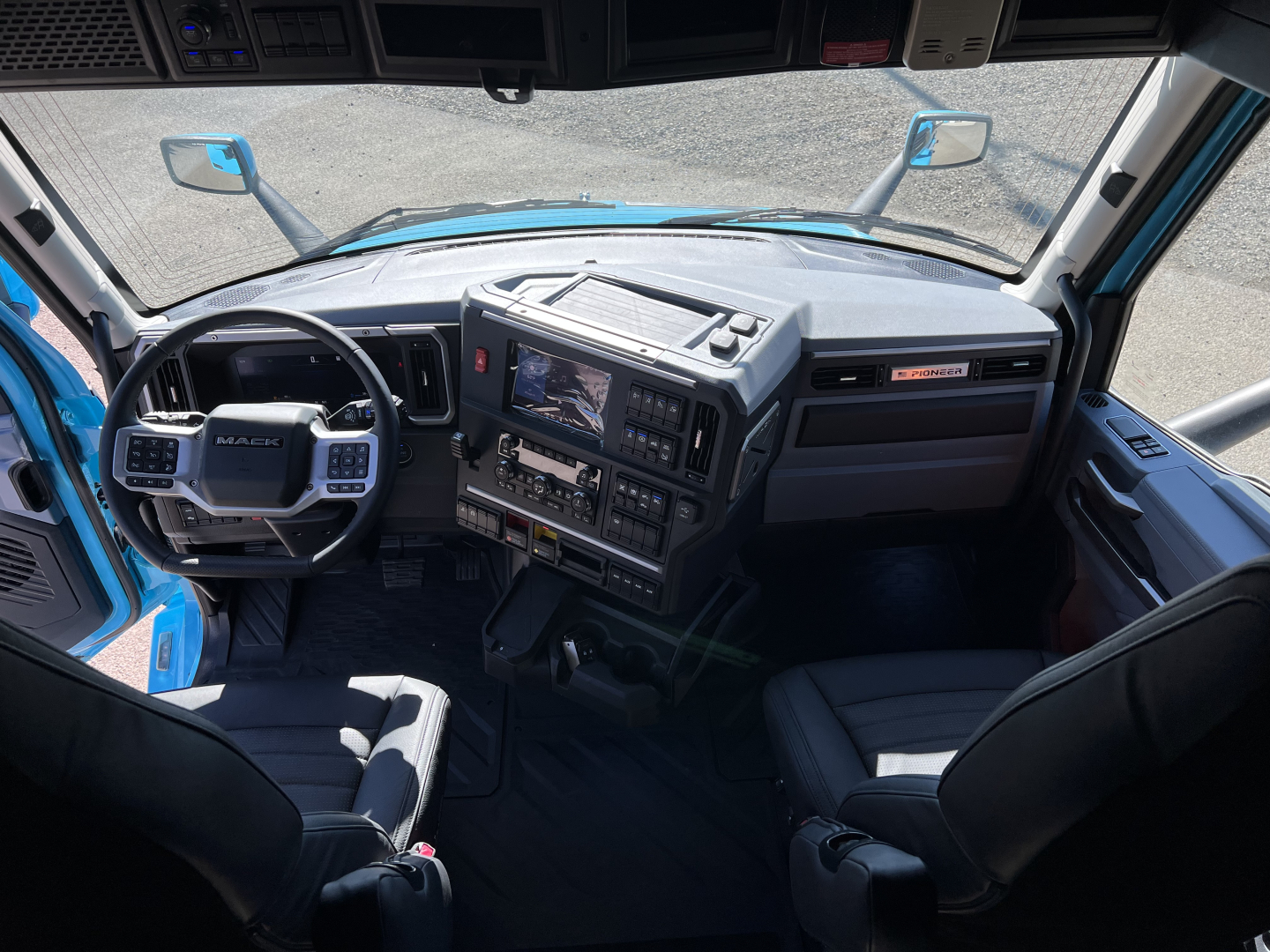 Interior of a Mack truck cab dashboard and steering wheel with many controls, large central screen, and driver and passenger seats visible.