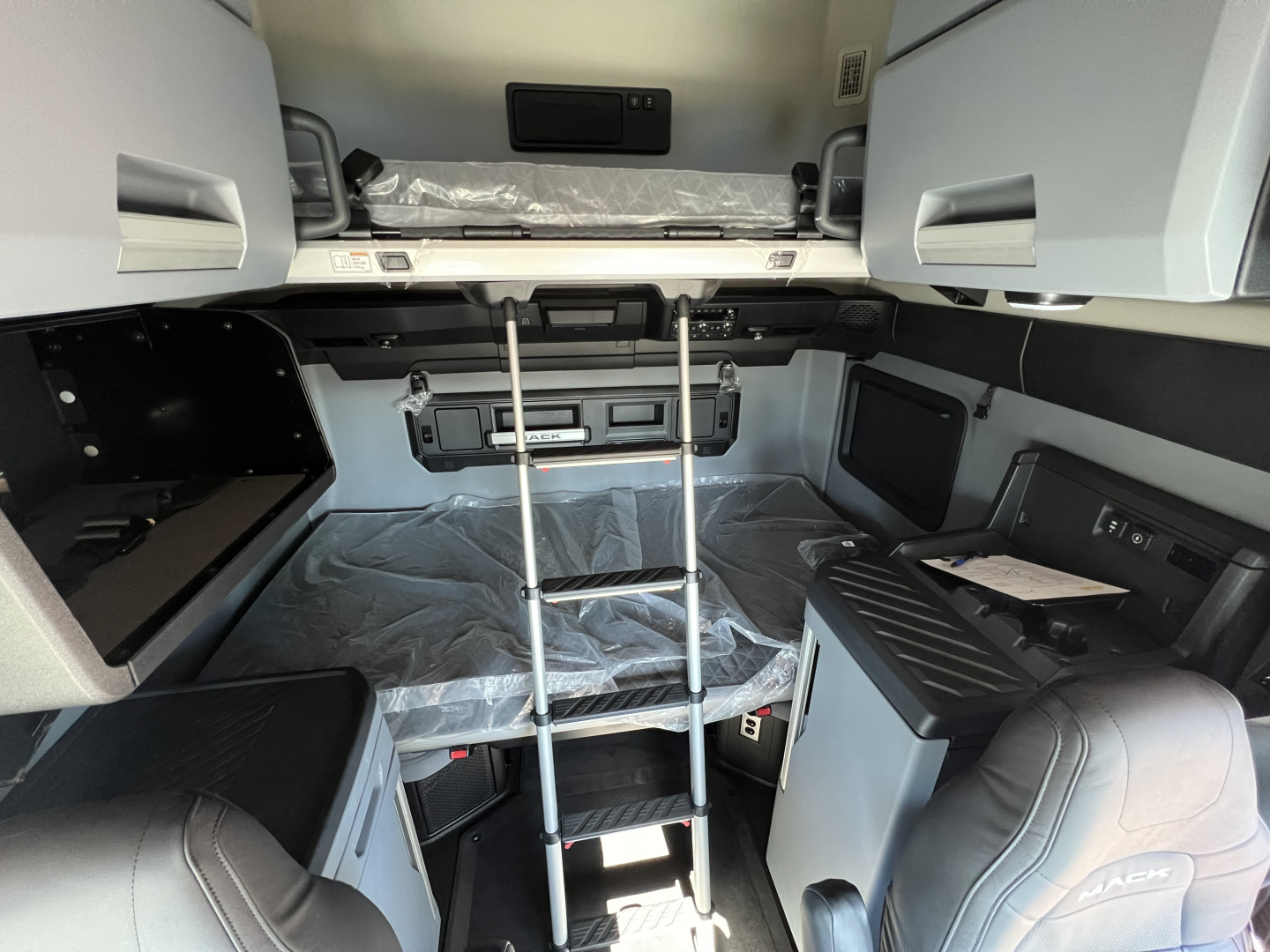 Truck cab interior showing bunk area with overhead storage, ladder, and side consoles in a new Mack PR64T76H sleeper model.