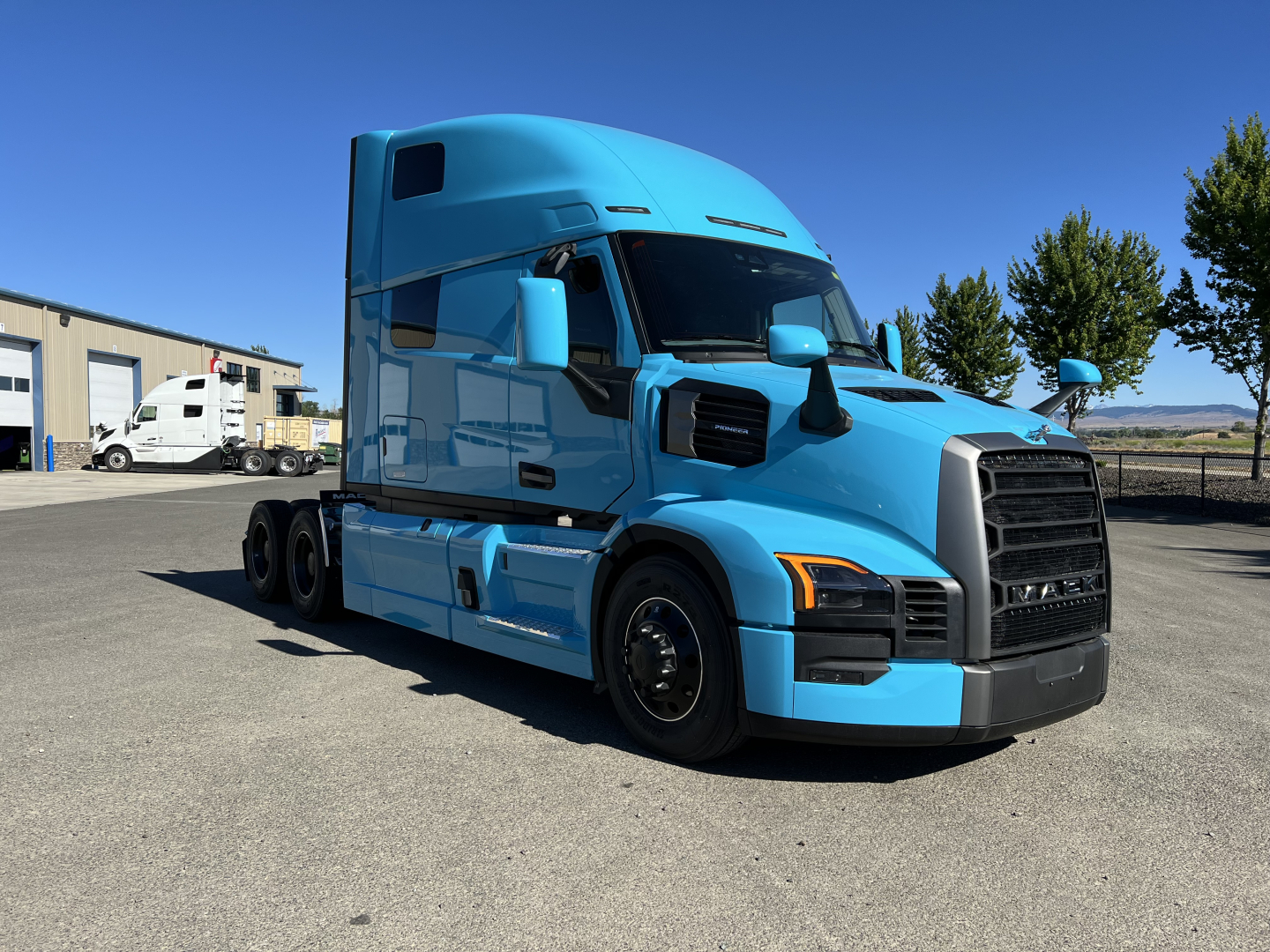 Blue Mack PR64T76H tractor-trailer cab in a parking lot, front three-quarter view, showing large grille, aerodynamic sleeper, and side mirrors.