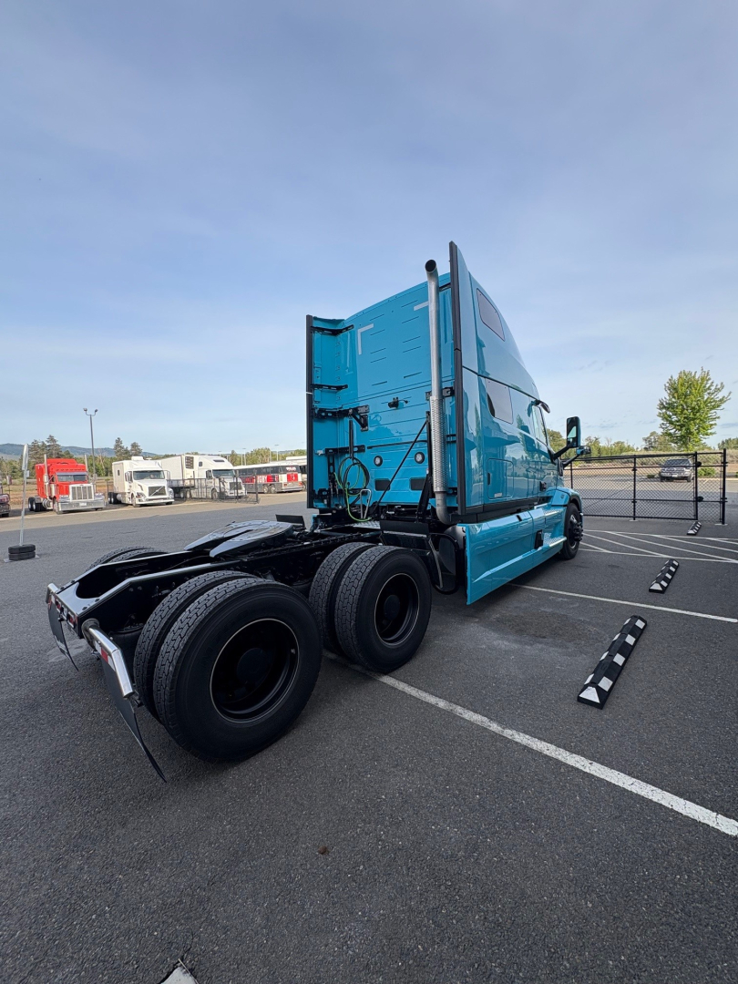Truck tractor rear three-quarter view showing blue cab, rear axle and fifth wheel with dual rear axles, no visible bumper plate; exterior has decals and tall exhaust stacks.