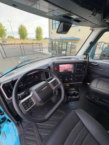 Truck cab interior showing Mack steering wheel, dashboard, and infotainment screen; blue exterior visible through windshield; driver seat and controls in view.