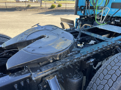 Truck chassis with large circular fuel/tank cover and exposed frame; visible hoses, cables, and wheel behind; blue cab in background; concrete lot.
