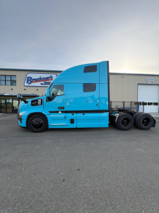 New blue Mack PR64T76H conventional sleeper truck side view, with two rear axles, parked in lot in front of dealership building.