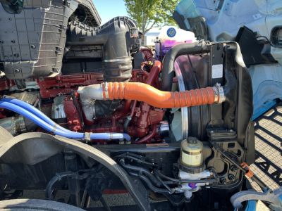 Mack PR64T76H engine bay showing orange radiator hose, blue hose, red engine block, power steering/auxiliary components and front mounted reservoir.
