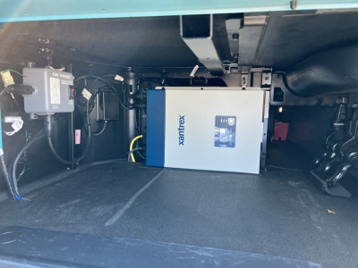 Truck cab interior under dash area showing Xantrex device in a blue box powered by wiring harnesses and a fuse block on left; toolbox or control box visible; floor area clean.