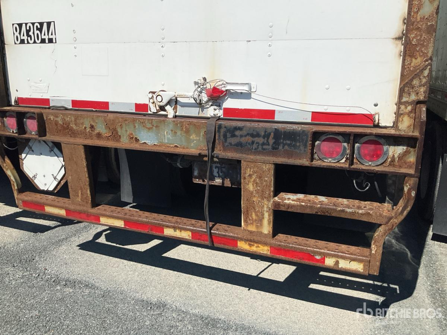 Rear of a trailer showing rusted metal frame and bumper with red circular taillights and a white panel container.