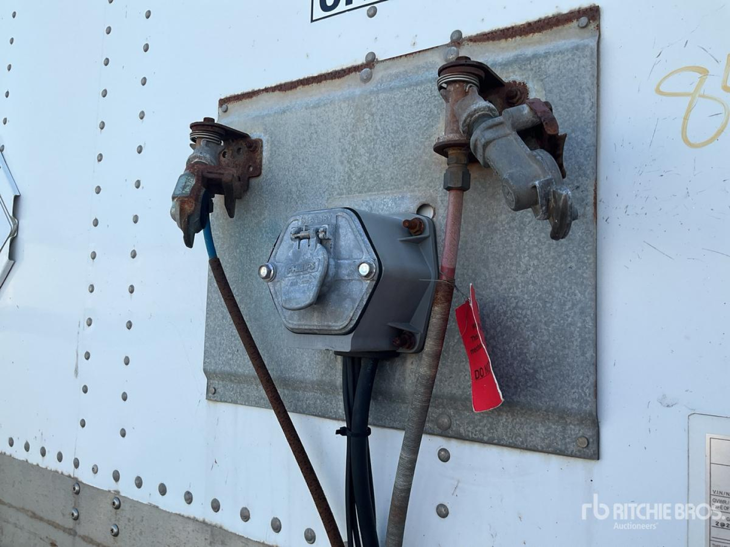 Trailer utility panel with two valve connections and a central electrical box mounted on a metal backplate, hoses and cables hanging, on a light-colored trailer wall.
