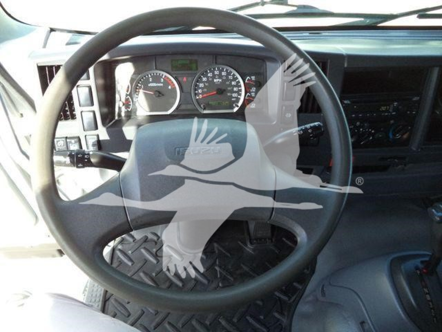 Isuzu truck interior steering wheel and dashboard dash showing gauges, pedals, and a diamond-plate floor mat.