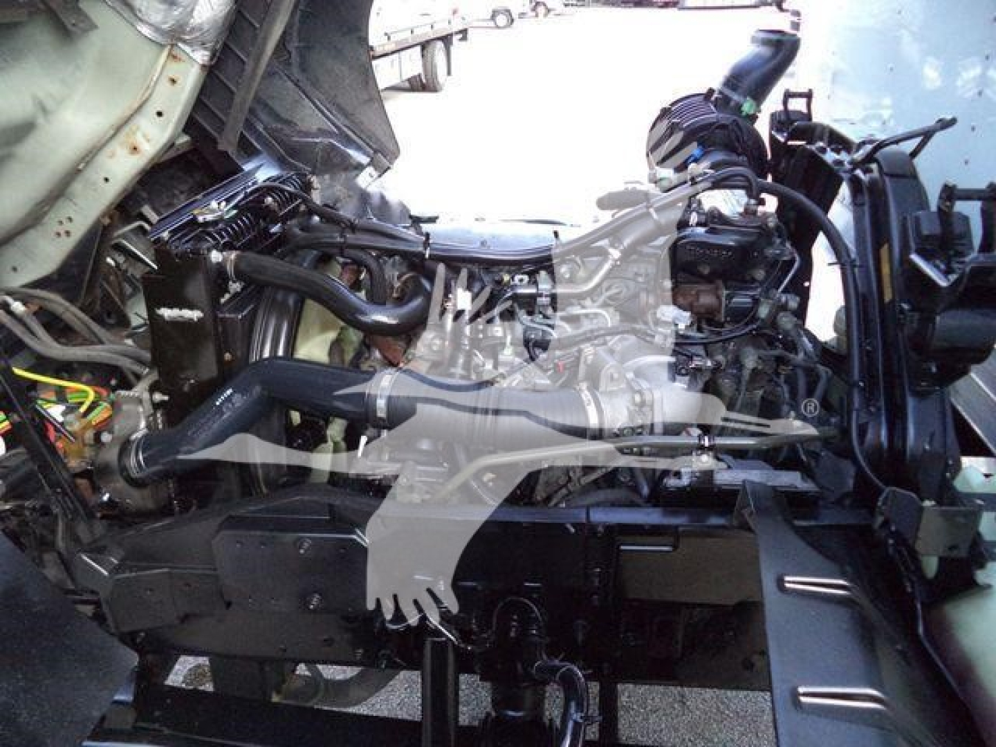 Isuzu FTR engine bay showing compact diesel engine with intake manifold, coolant reservoir, and assorted hoses in a truck front section.