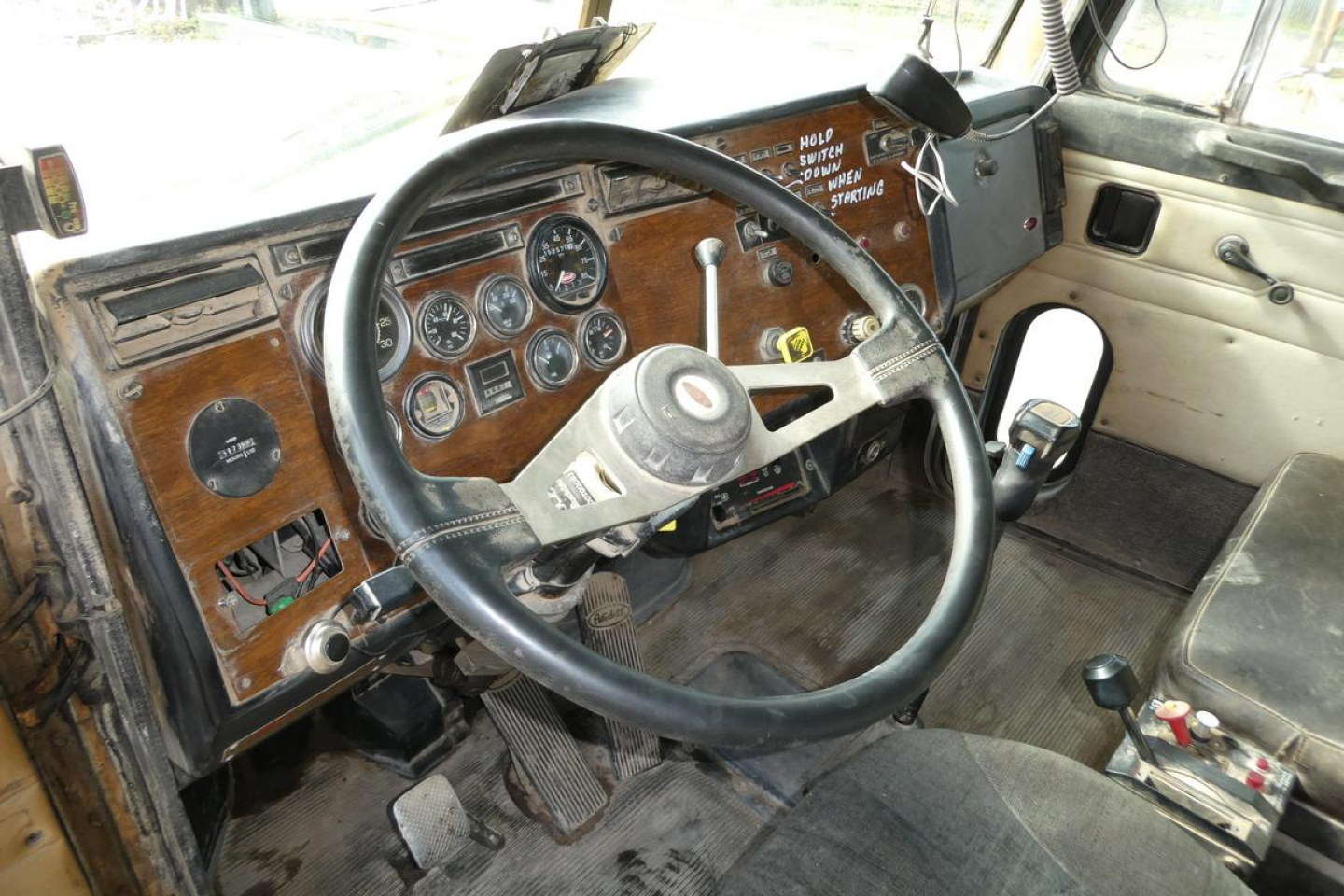 Peterbilt truck cabin dashboard with wood paneling and analog gauges, steering wheel, gear shift, and driver's seat in view.