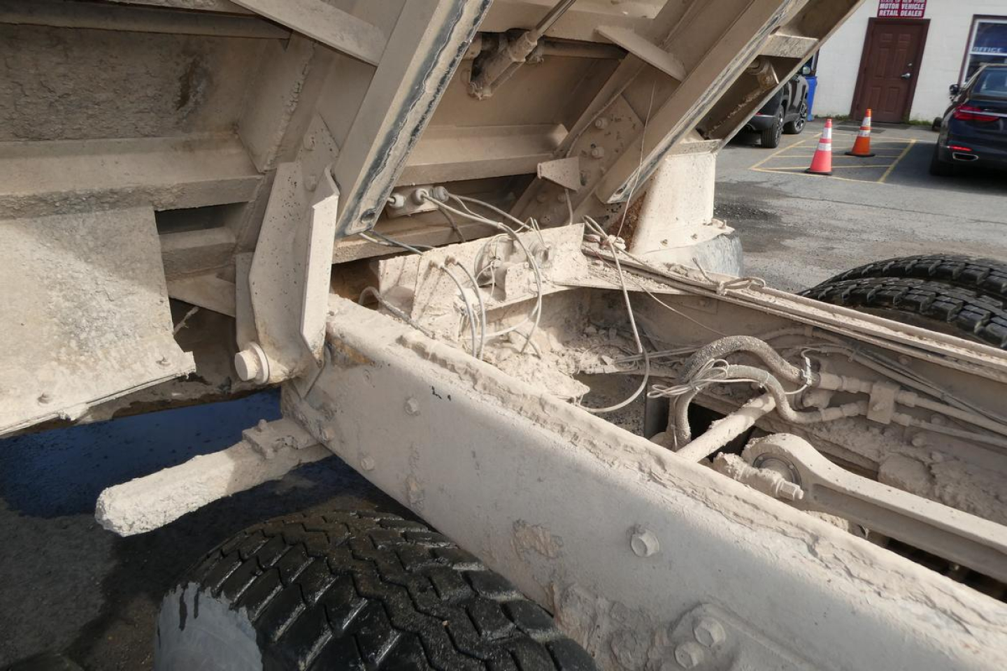 Truck chassis underbody with dirt-covered frame and suspension, visible bolts, wiring, and hydraulic hoses around the dump bed hinge area.