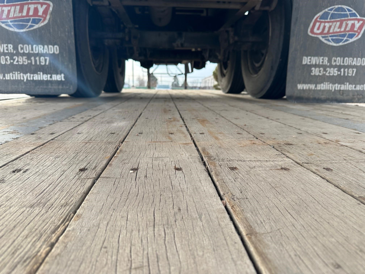 Trailer underside view showing wooden deck boards between dual rear axles; mud flaps with Utility logo and Denver, Colorado contact info visible.