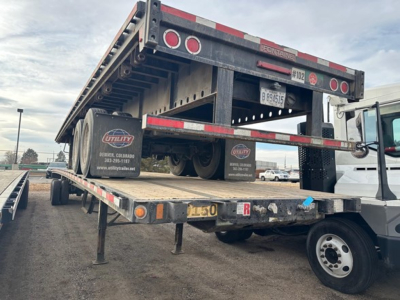 Flatbed trailer in a raised position showing the bed and rear wheels with utilities decals and license plate framing. Side rails, rear lights, and support legs visible.
