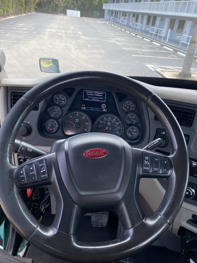 Peterbilt steering wheel in-cabin view showing dashboard gauges and a central display screen with warning/notification text. Brand logo on the wheel.
