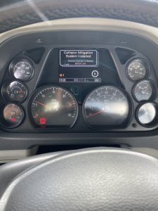 Peterbilt 579 instrument cluster with central display showing Collision Mitigation System installed message, surrounded by gauges including RPM, speed, and fuel. Steering wheel and dashboard visible.