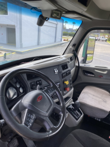 Peterbilt 579 truck interior showing steering wheel with Peterbilt emblem, dash gauges, center console, and driver's seat. Windshield with sun visor, side window, and rearview mirror visible.
