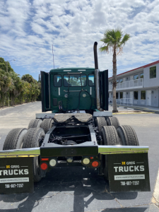 Peterbilt 579 rear view showing dual rear axles and green cab, with mud flaps reading Greg Trucks in a dealership lot.