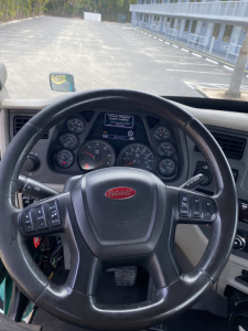 Peterbilt steering wheel in-cabin view showing dashboard gauges and a central display screen with warning/notification text. Brand logo on the wheel.