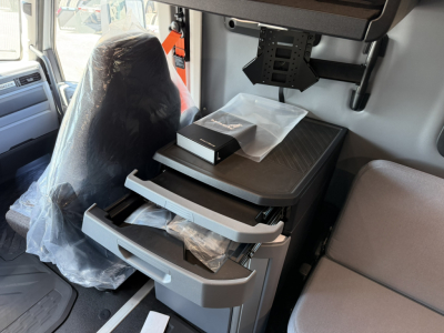 Cab interior of 2027 Mack PR64T76H showing driver seat, center console with stacked drawers and a small black box on top, plastic coverings.