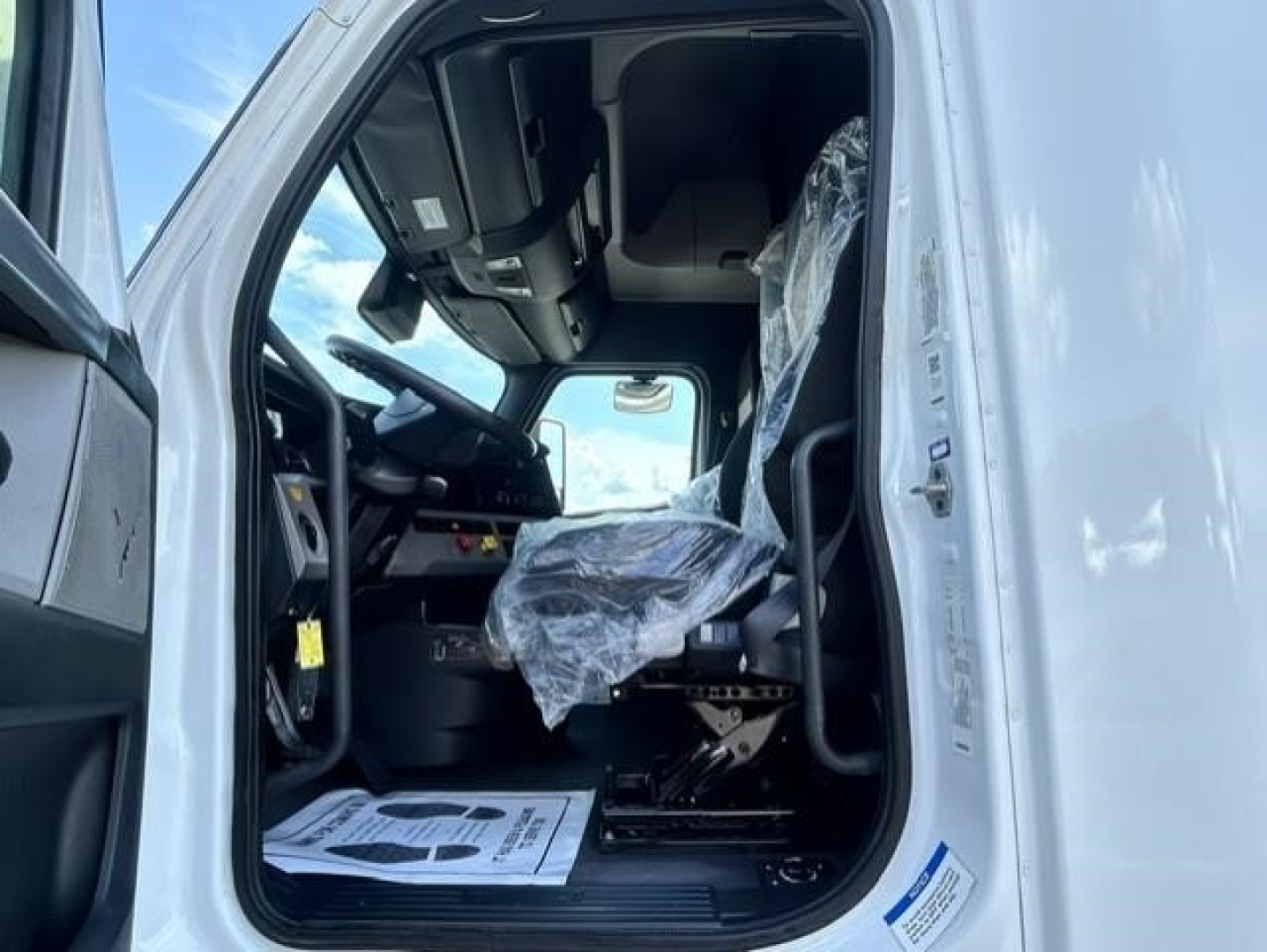 Freightliner Cascadia 126 truck cab interior view showing driver's seat with plastic cover, steering wheel, dashboard, and entry door frame.