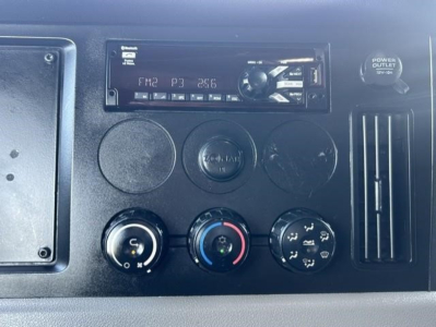 Dashboard panel of a Freightliner Cascadia showing radio unit, circular knobs for climate control, and a power outlet along with air vent.