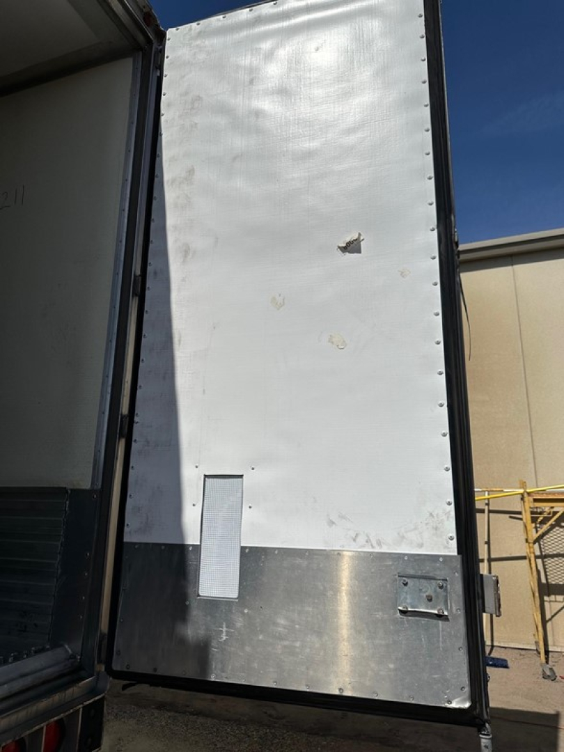 Front corner of a refrigerated trailer with a metal lower panel and white insulated upper door. Minor dents and scuffs on the white surface; small rectangular vent and metal latch visible on the lower panel.