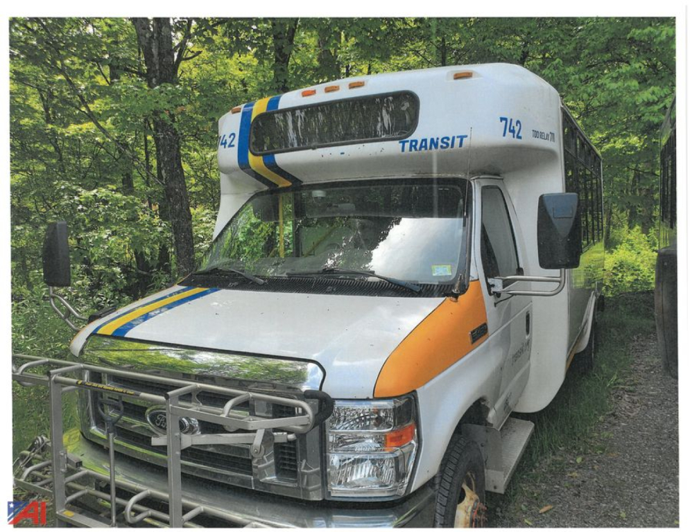 Ford E450 bus front-left view showing grille guard and blue Transit markings, orange stripe on white body, wooded background.