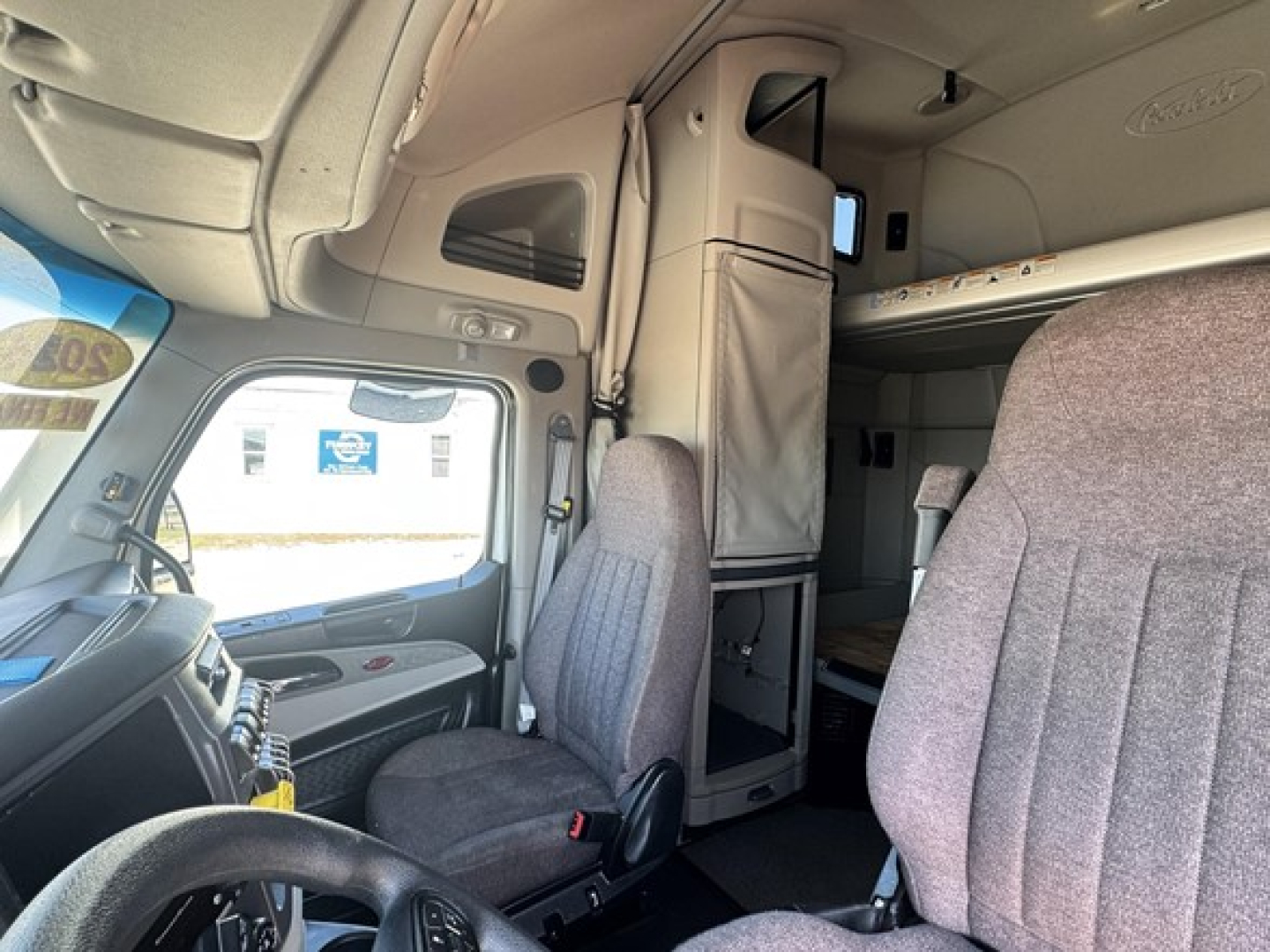 Truck cab interior: driver seat and passenger seat with overhead sleeper berth area, dashboard and window visible.