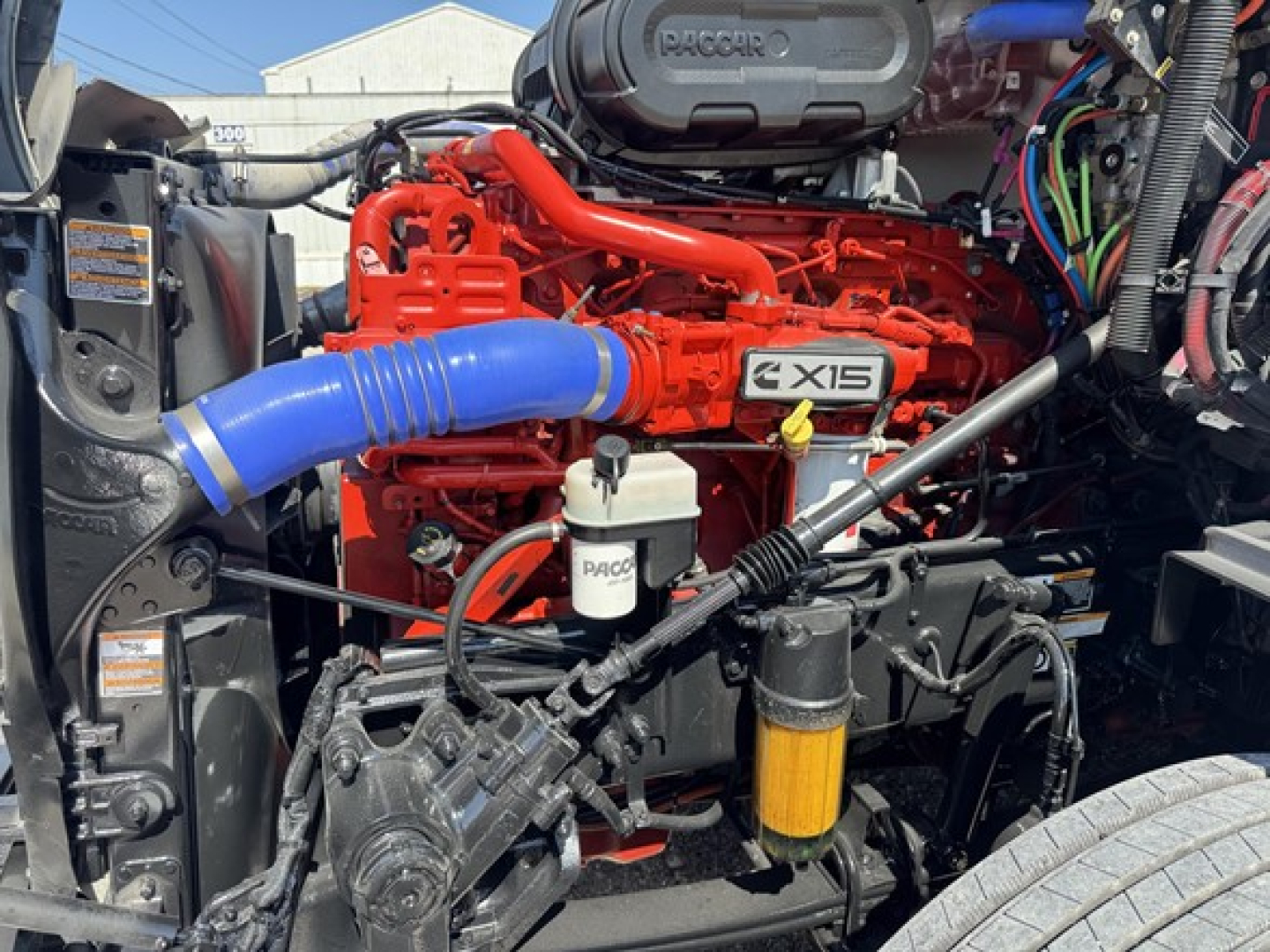 Peterbilt 579 engine bay showing red Cummins X15 engine with blue intercooler hose, white reservoir, and various black hoses and wiring.