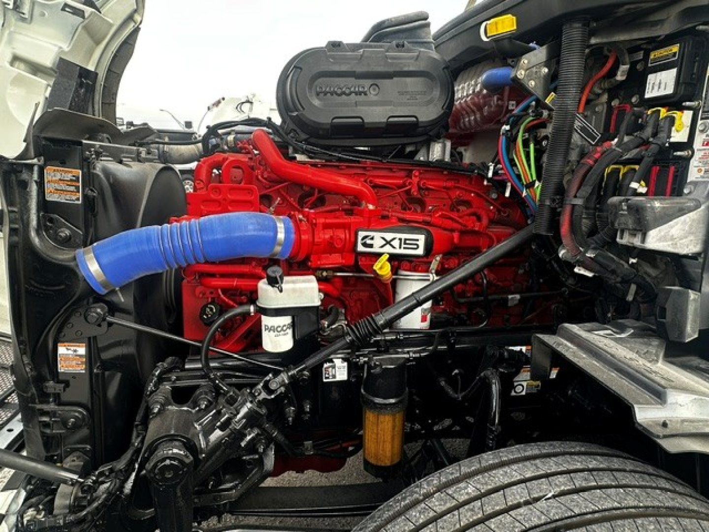Peterbilt 579 engine bay showing red Cummins engine with blue hose, black air intake cover, and various wiring harnesses.