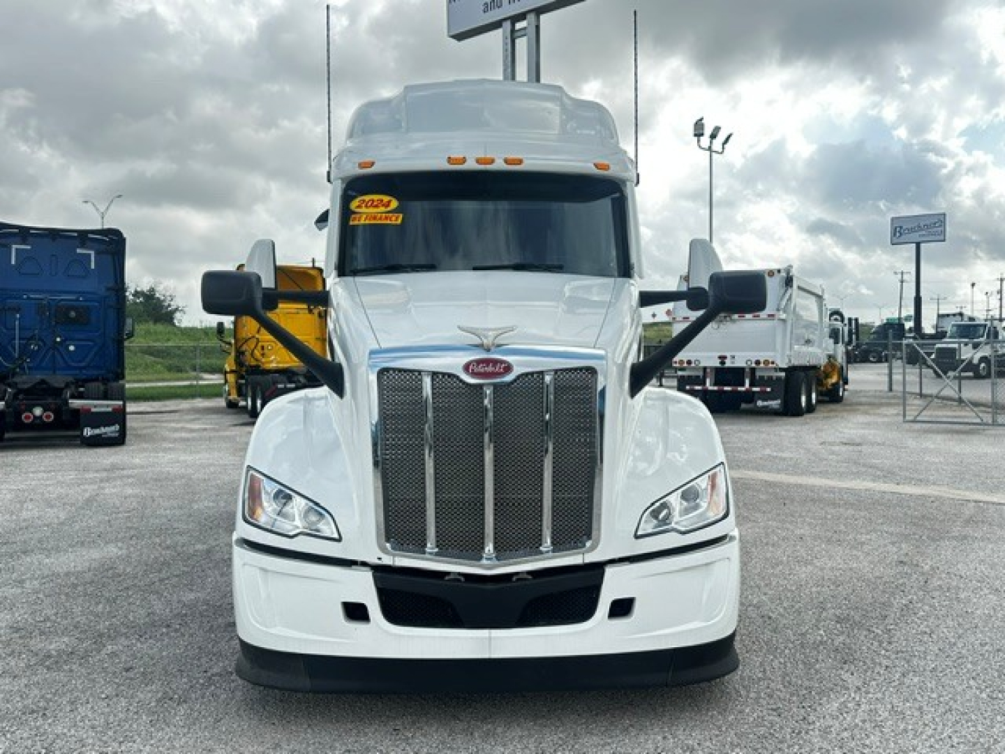 Peterbilt 579 front view in white, with large chrome grille and bumper; cab and mirrors visible; yellow and white trucks in background.