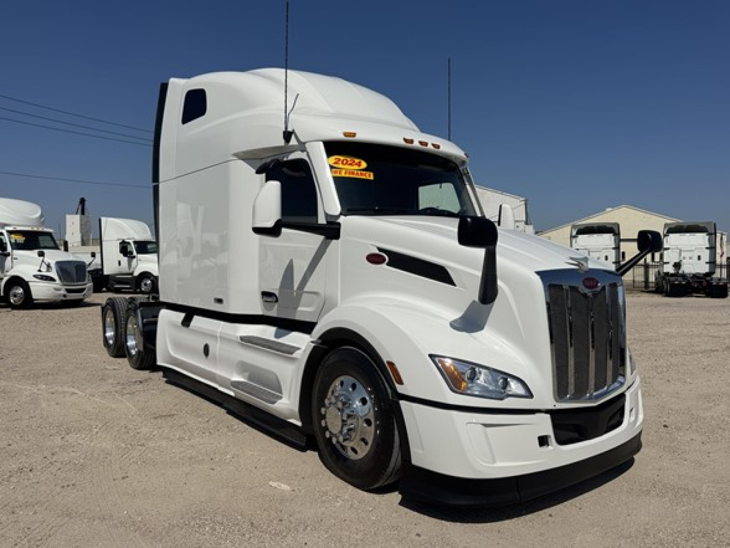 Peterbilt 579 sleeper cab tractor, white exterior, front chrome grille, yellow 2024 sticker on windshield, side view showing front wheels and exhaust area.