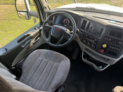 Peterbilt 579 cab interior showing steering wheel, dashboard with instrument cluster, and center console. Gray fabric seat, controls on door and dash, side mirror visible.