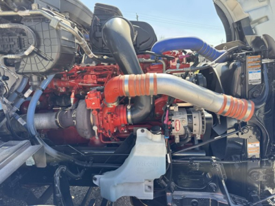 Peterbilt 579 engine bay showing red engine block, large intake piping, turbo, coolant hoses, and auxiliary components.