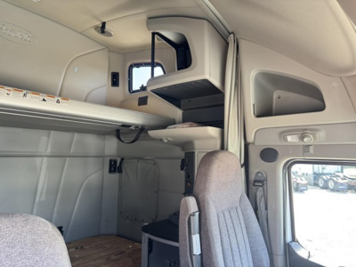 Peterbilt sleeper cab interior showing overhead storage shelf and dashboard area with window; gray upholstery and cab components visible.