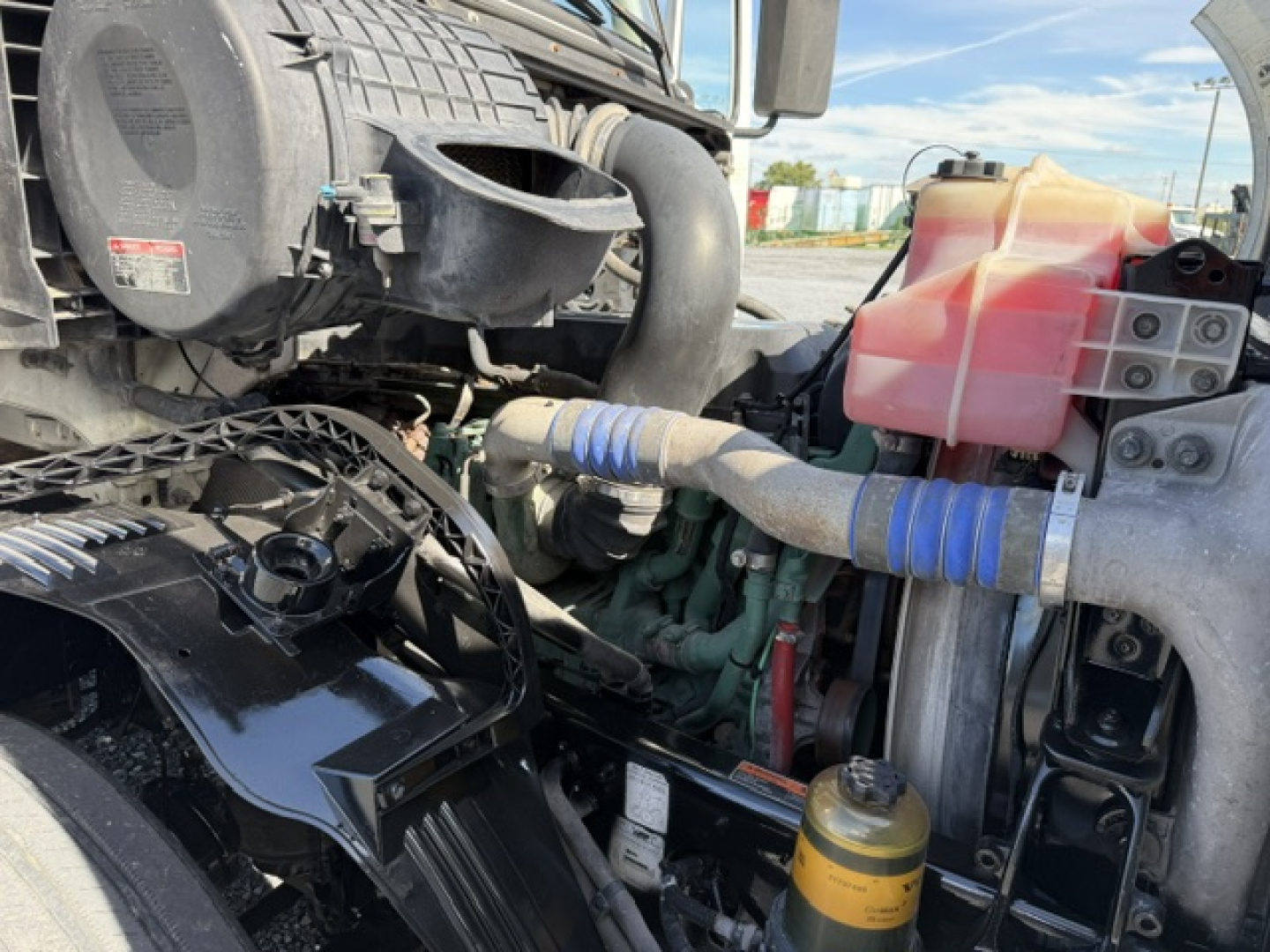 Truck engine bay of a 2016 Volvo VNL64T300 showing coolant reservoir, radiator and hoses, blue hose clamps, yellow oil fill cap, and pink/red coolant reservoir.