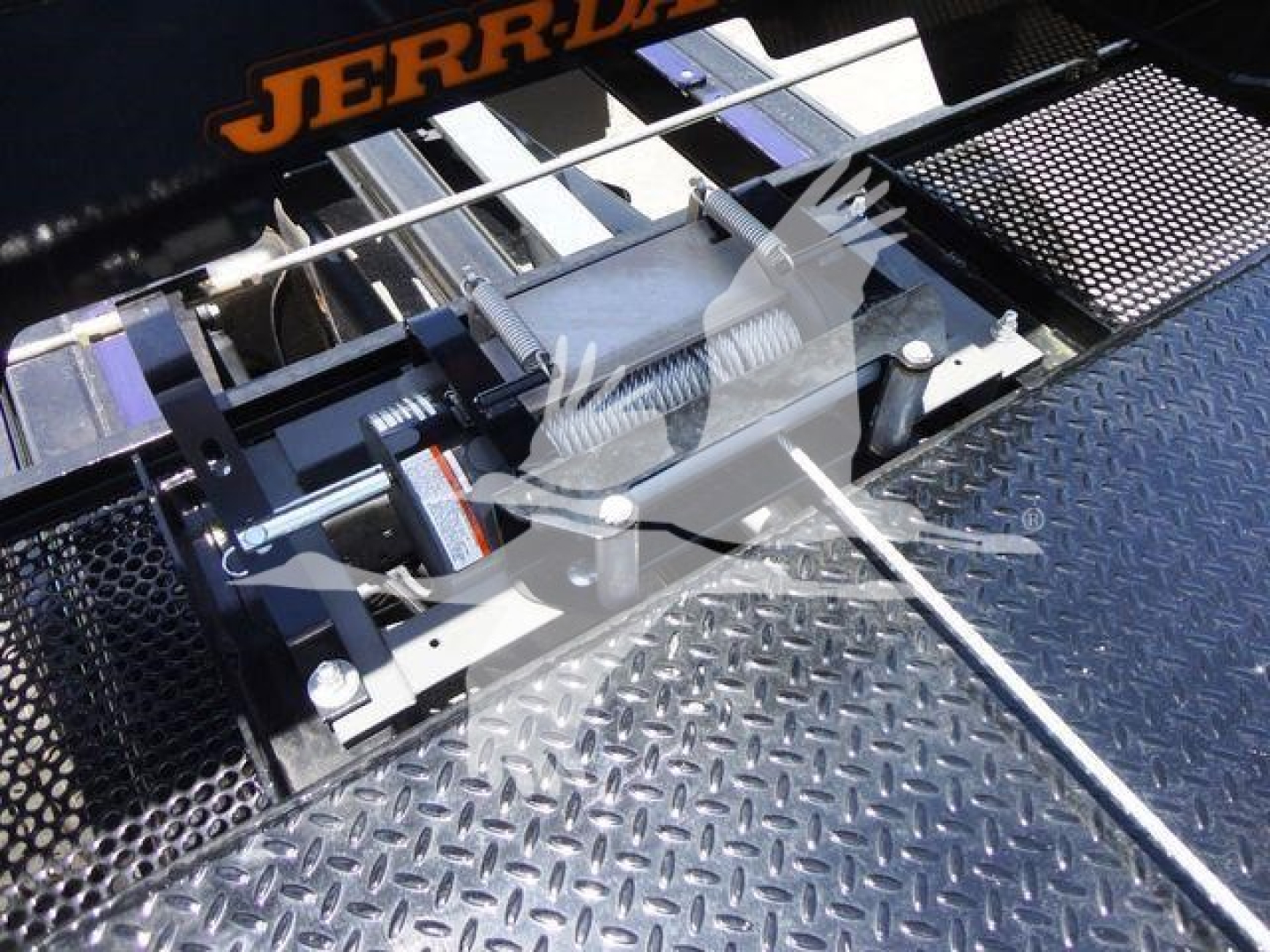 Tow truck rollback mechanism with metal winch and roller shaft, hydraulic line visible, diamond plate surface, label JERR-DYNE on top rail.