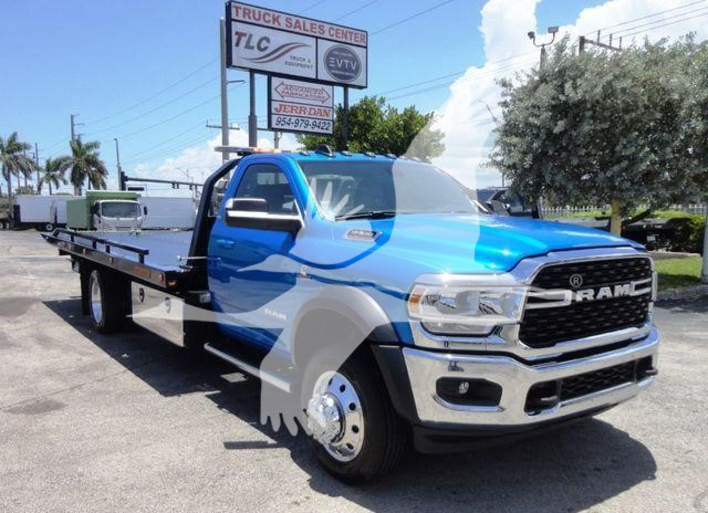 Blue Ram 5500 rollback tow truck with flatbed, front grille shows RAM emblem, dual rear wheels visible, dealership lot in background.