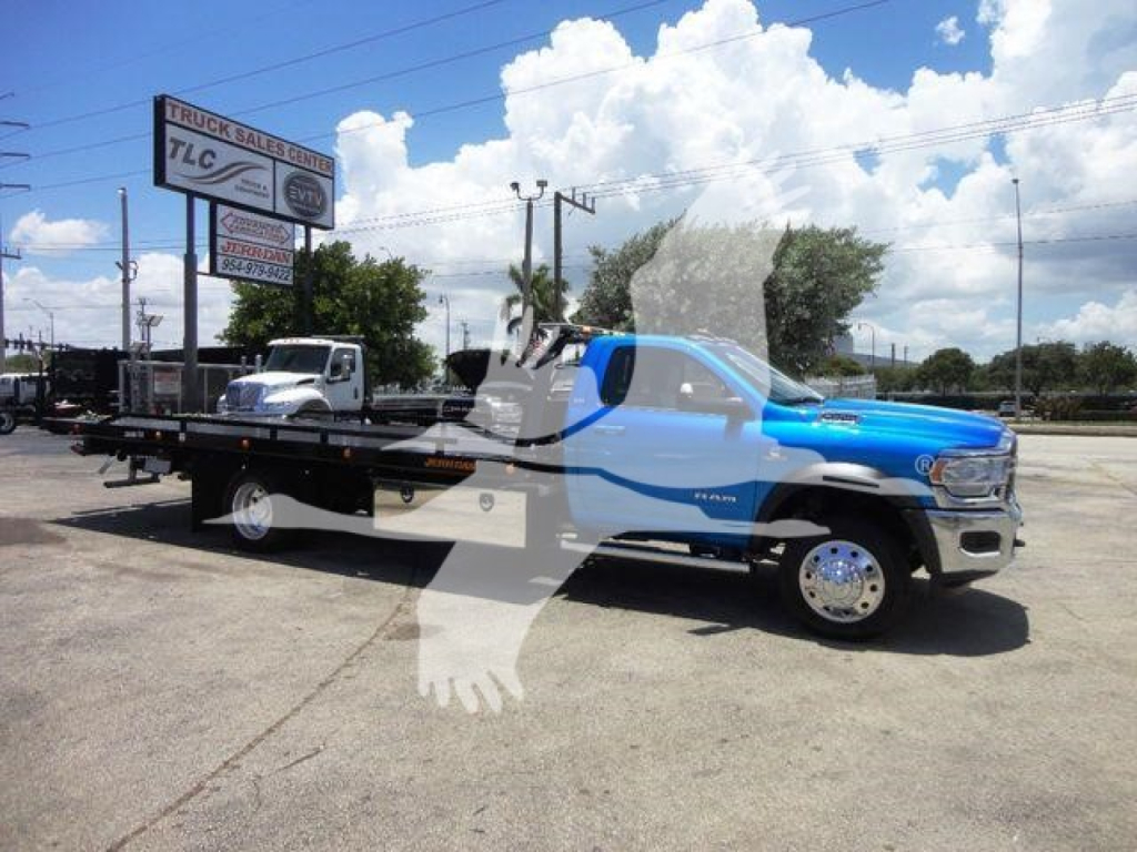 Tow truck with a blue and white Ram 5500 SLT flatbed rollback trailer in a lot, stock number J33507, category Rollback, new truck.