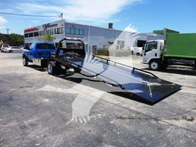 Ram 5500 rollback tow truck with a long flatbed in a dealership lot, blue cab, white building in background.