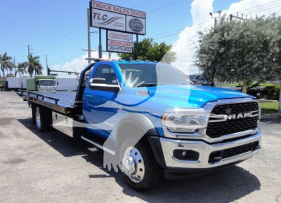 Blue Ram 5500 rollback tow truck with flatbed, front grille shows RAM emblem, dual rear wheels visible, dealership lot in background.